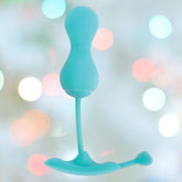 A turquoise OhMiBod Lovelife Krush Smart Kegel Fitness by OhMiBod, featuring a curved shape with a bulbous top and thin stem, is set against a softly blurred background with multicolored circular lights.